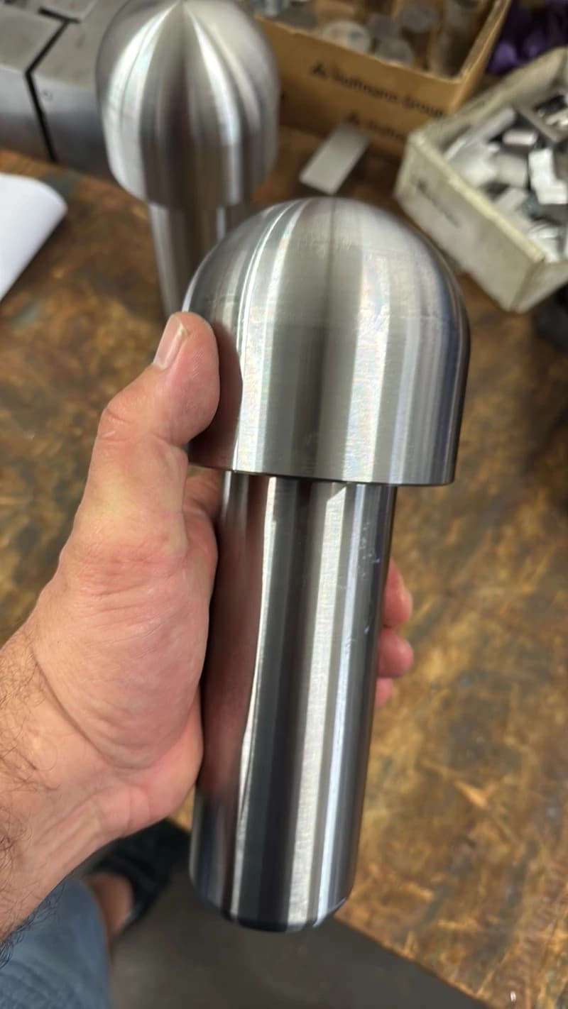 Eine Hand hält ein poliertes Metallobjekt in Form eines Pilzes. Im Hintergrund sind weitere Metallobjekte und Werkzeugkisten zu sehen.