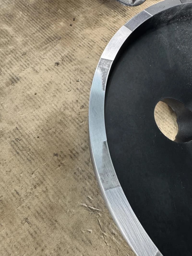 Nahaufnahme eines schwarzen Metallrads mit einem silbernen Rand auf einer rauen Oberfläche. Das Rad hat eine zentrale Öffnung und der silberne Rand ist in Abschnitte unterteilt.