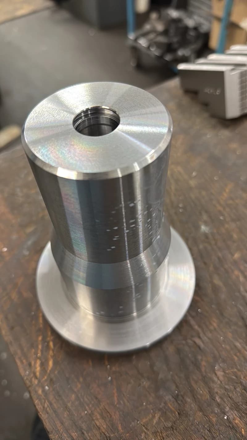 Nahaufnahme eines bearbeiteten Metallteils mit einem Gewindeloch auf einem Holztisch. Das Teil scheint auf einer Drehmaschine bearbeitet worden zu sein und weist eine glatte, reflektierende Oberfläche auf.