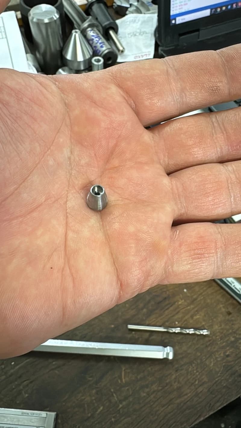 Ein kleines, metallisches Kegelteil liegt in einer Hand. Im Hintergrund sind Werkzeuge und ein Laptop zu sehen.