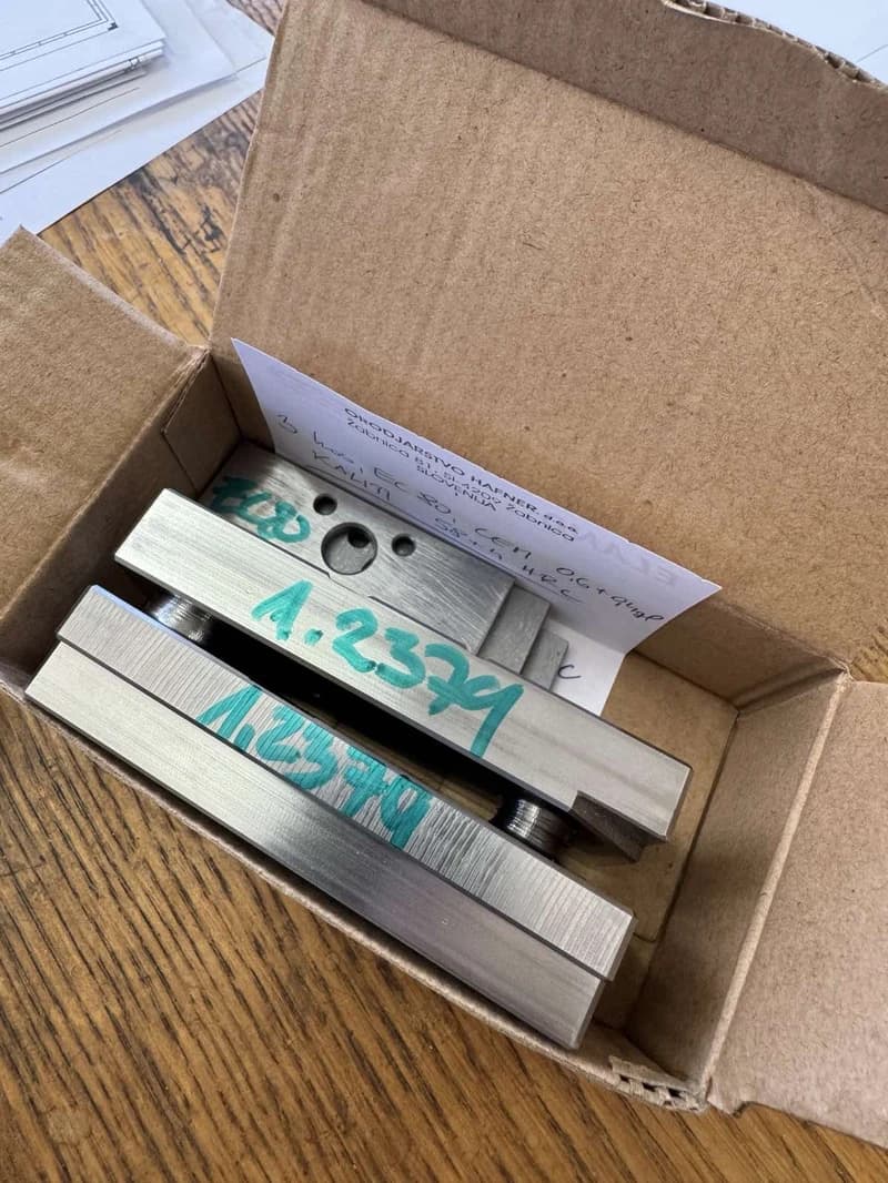 Metallteile mit der Aufschrift "1.2379" liegen in einer Pappschachtel, zusammen mit einem Zettel mit der Aufschrift "ORODJARSTVO HAFNER". Die Teile scheinen maschinell bearbeitet zu sein.