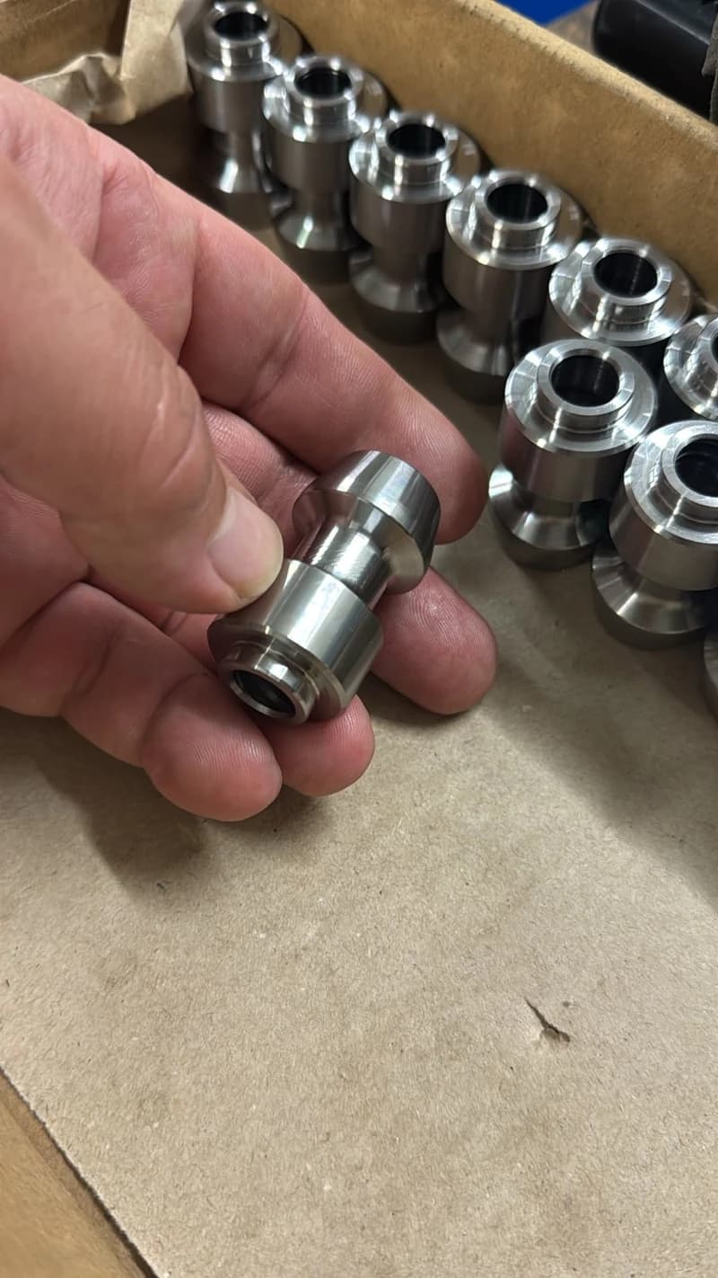 Einige metallische, spindelförmige Teile in einer Pappschachtel, wobei eines von einer Hand gehalten wird. Die Teile scheinen neu gefertigt und bereit für die Inspektion zu sein.