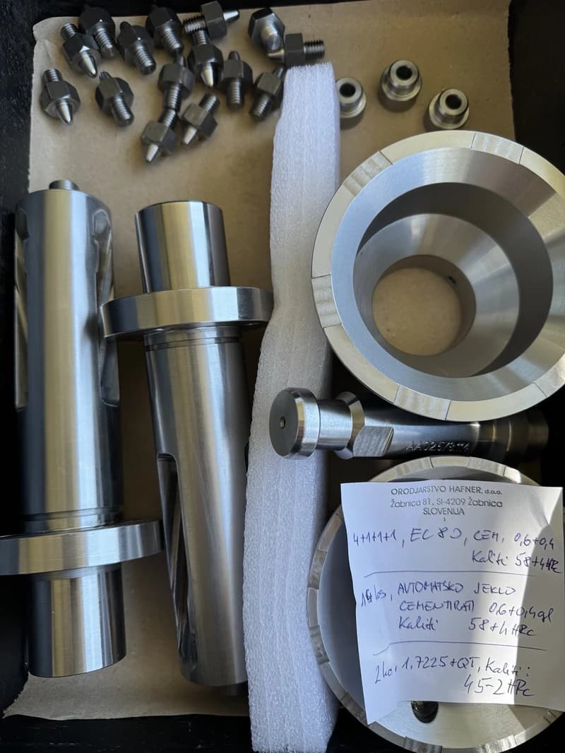 Ein Tablett mit verschiedenen Metallteilen, darunter Schrauben, Hülsen und zylindrische Werkzeuge, zusammen mit einem Zettel mit dem Firmennamen Orodjarstvo Hafner aus Slowenien.