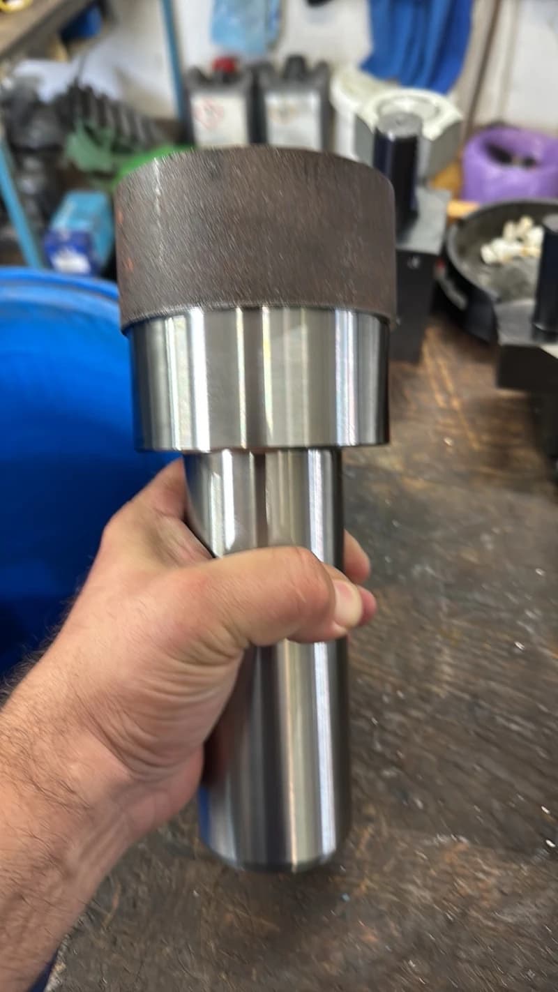 Ein Mann hält einen zylindrischen Metallgegenstand mit einem braunen, zylindrischen Aufsatz in der Hand. Der Gegenstand scheint ein Werkzeug oder ein Maschinenteil zu sein.