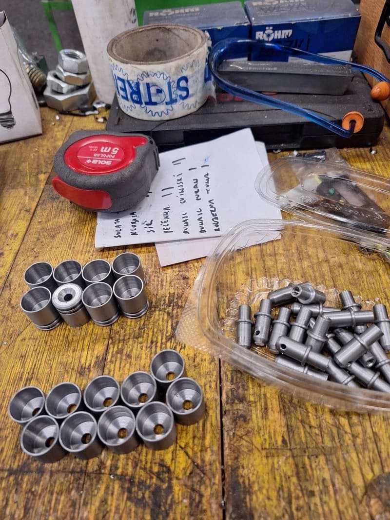 Auf einer Werkbank liegen verschiedene Metallteile, ein rotes Messband und ein Blatt Papier mit Notizen. Die Metallteile sind in Gruppen angeordnet und scheinen zu einem größeren Projekt zu gehören.